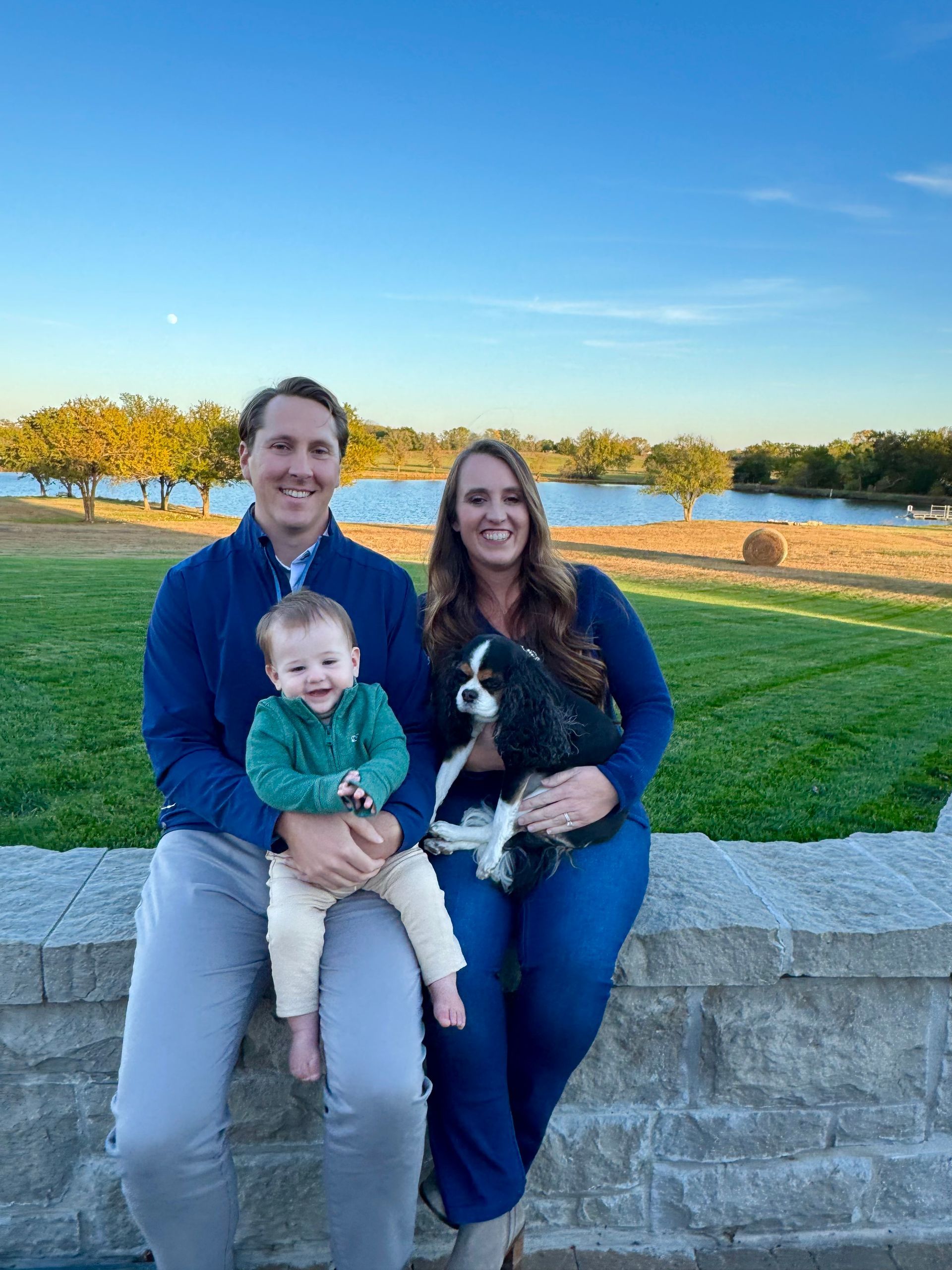 A family sits on a stone wall outdoors with a small dog, posing with a smiling toddler against a lake and blue sky.