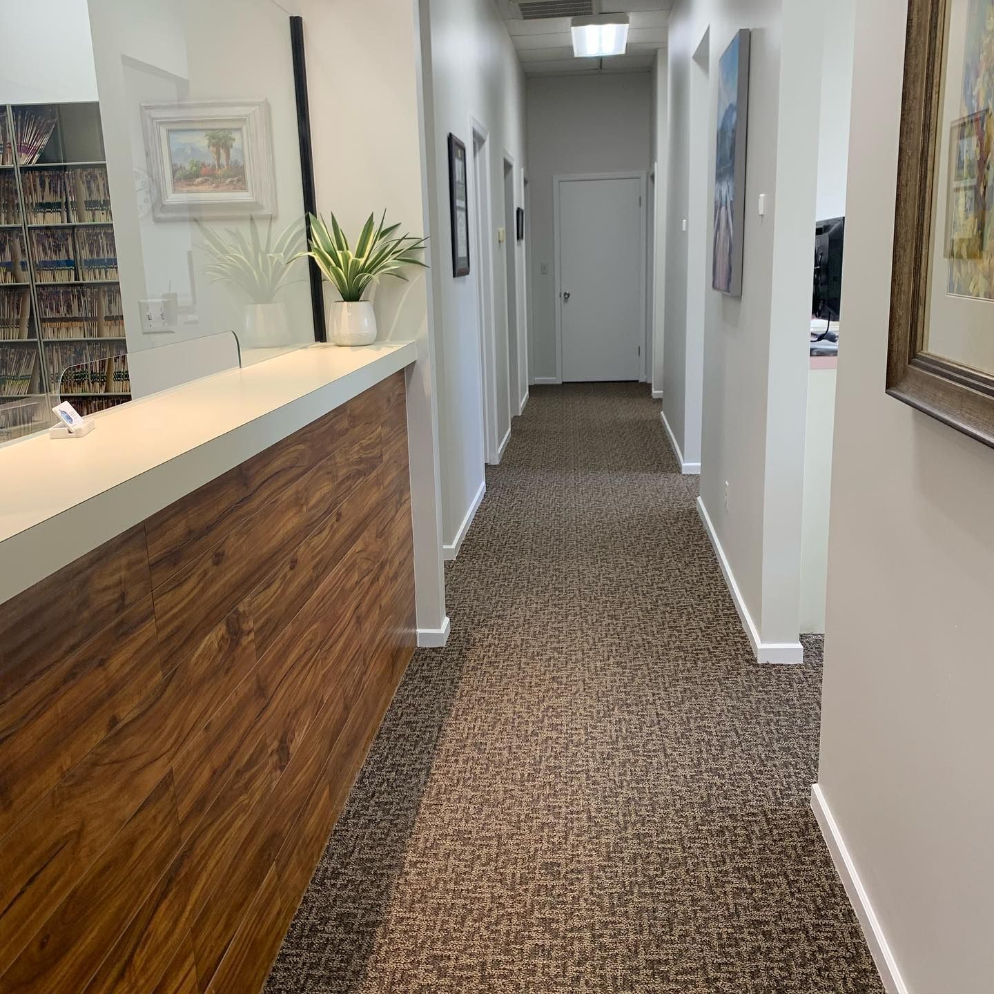 A reception desk with a wood-grain front faces a carpeted hallway leading to a white door in a professional office.