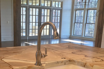 Kitchen faucet on a light-colored countertop, with large windows and wooden trim in the background.