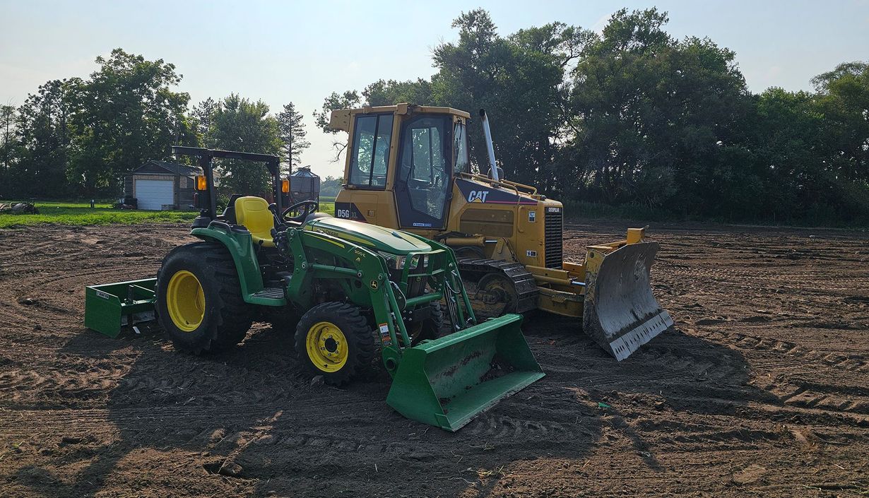 Green tractor and yellow bulldozer parked in a dirt field under trees.