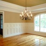 Empty dining room with wood floors, wainscoting, and a chandelier. Large arched window provides outside view, gold ceiling, beige walls.