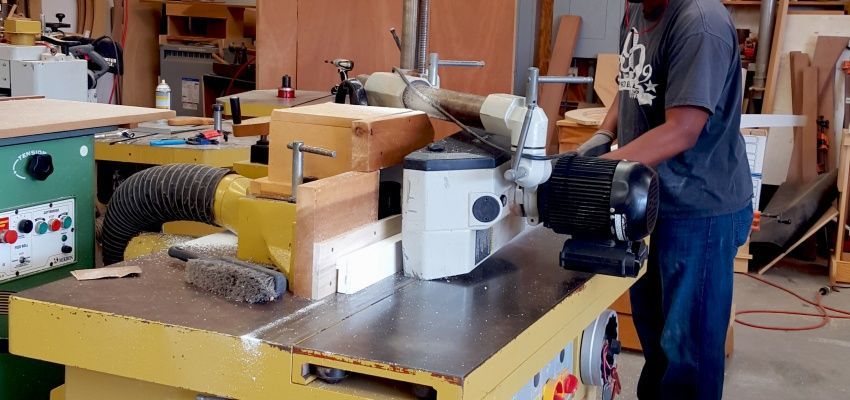 A person operates a woodworking machine in a workshop. The machine is yellow, with the person facing it and holding the handle.