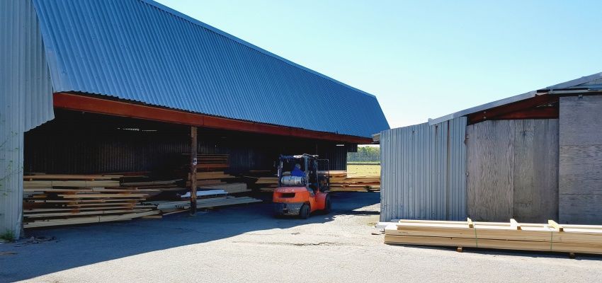 A forklift moves lumber at a lumber yard. Blue roofed structures are in the background.