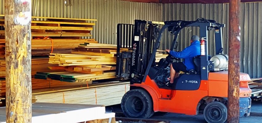 A worker operates an orange forklift inside a lumberyard, loading wooden beams. He wears a blue jacket.