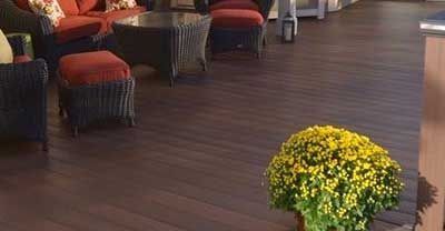 Dark brown wooden deck with outdoor wicker furniture, including a sofa and ottoman, with red cushions. A yellow flowering plant is in the foreground.