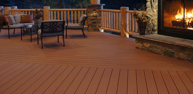 Outdoor deck with seating area and fireplace. Fireplace has a burning fire. Brown deck with a brown and stone fireplace.