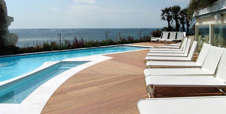 A luxury pool and deck with white lounge chairs overlook the ocean on a sunny day.