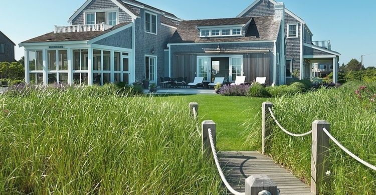 Coastal home with gray siding and a grassy lawn. Wooden walkway leads through tall grass.