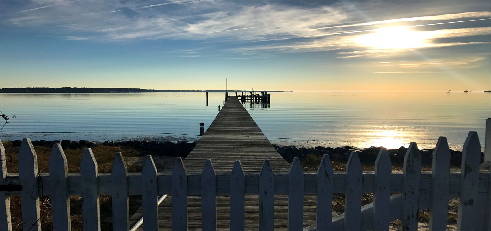 Wooden pier extending into calm water under a blue sky and sunlight. A white picket fence is in the foreground.