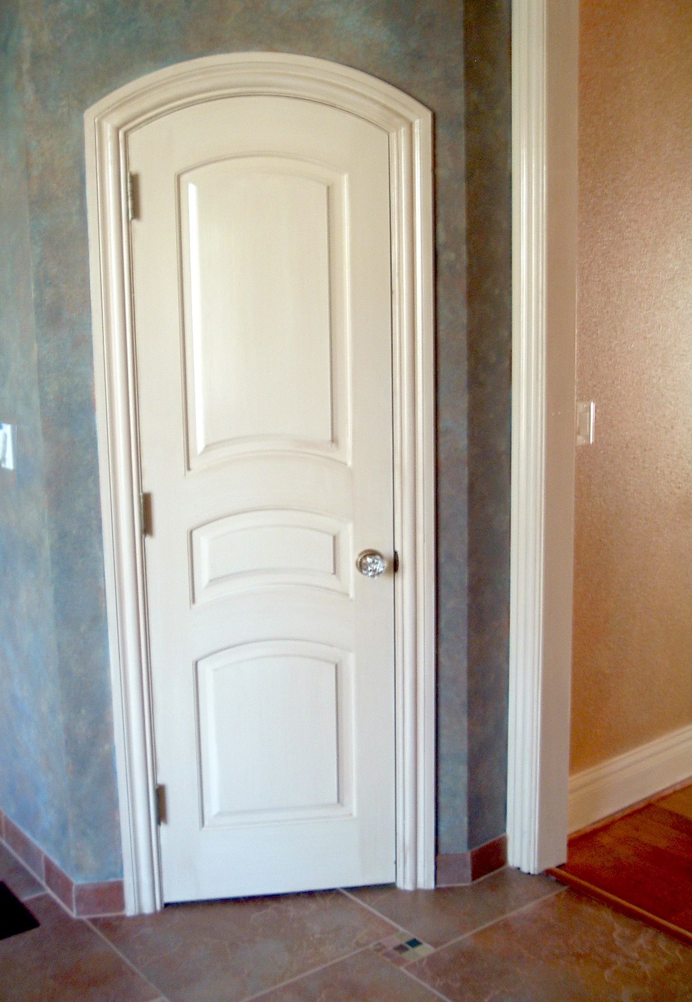 White, arched-top door in a beige and blue-gray hallway. Door has panel detailing with a crystal doorknob.
