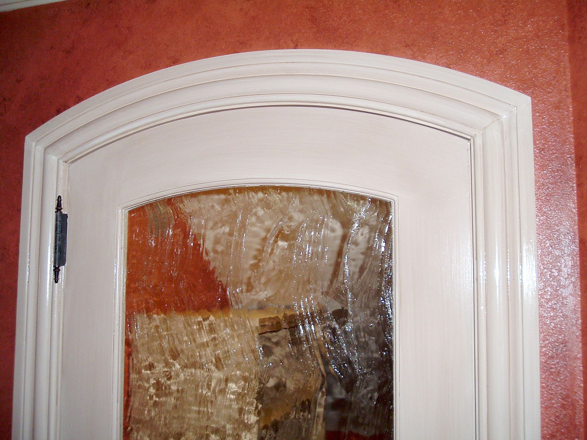 Cream-colored cabinet with arched glass door, set in a reddish-brown wall. The cabinet has a dark hinge.
