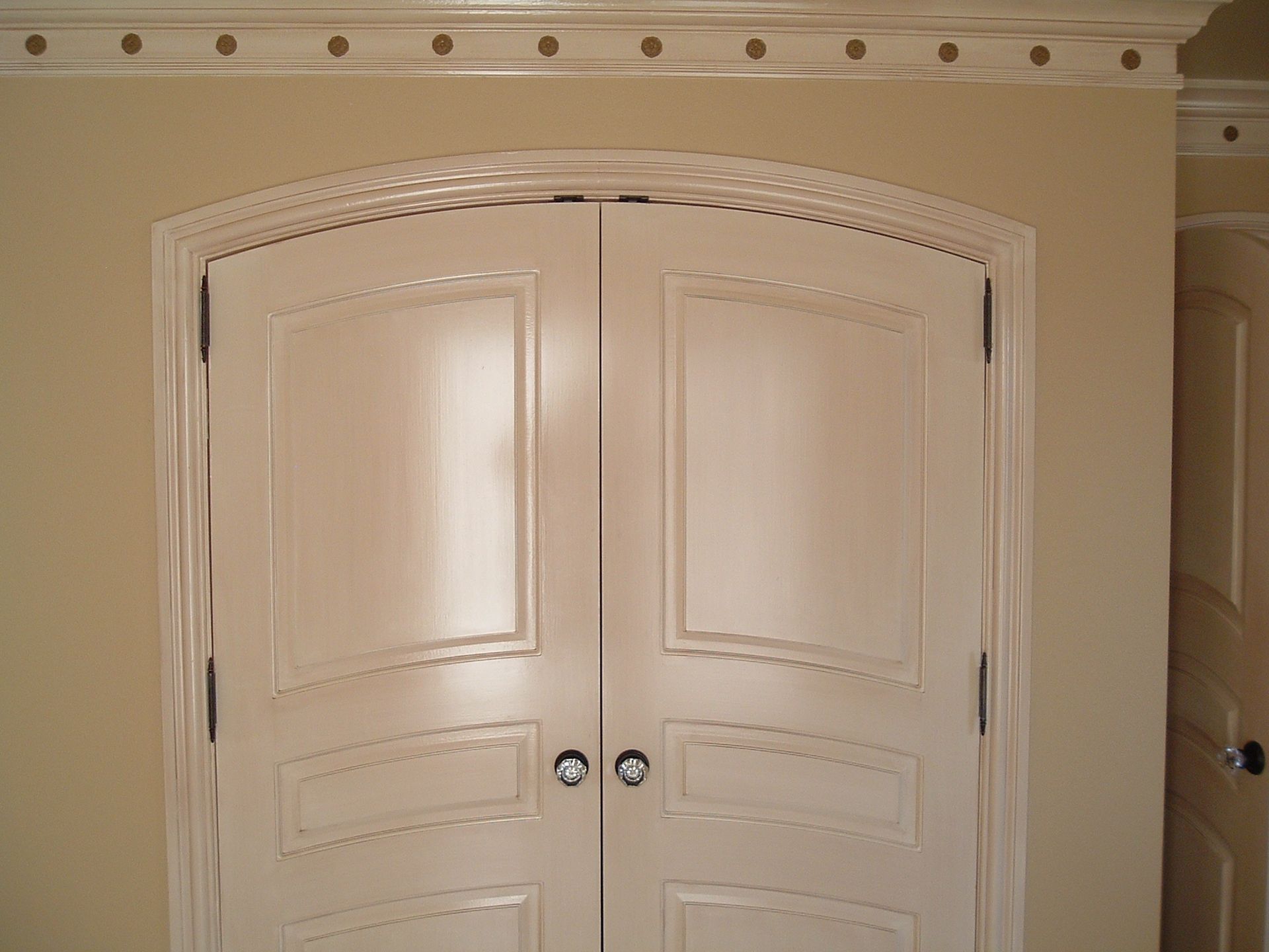Double doors with arched panels and dark hardware, painted off-white, set against a beige wall.