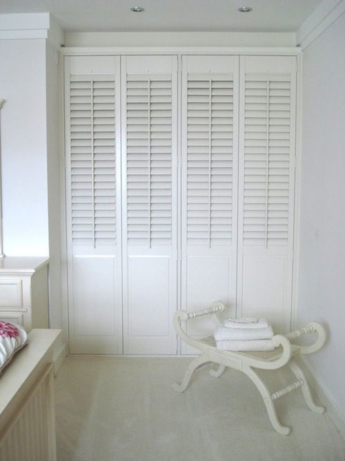 White closet doors with shutters, closed, next to a white bench with folded towels on a white carpet.
