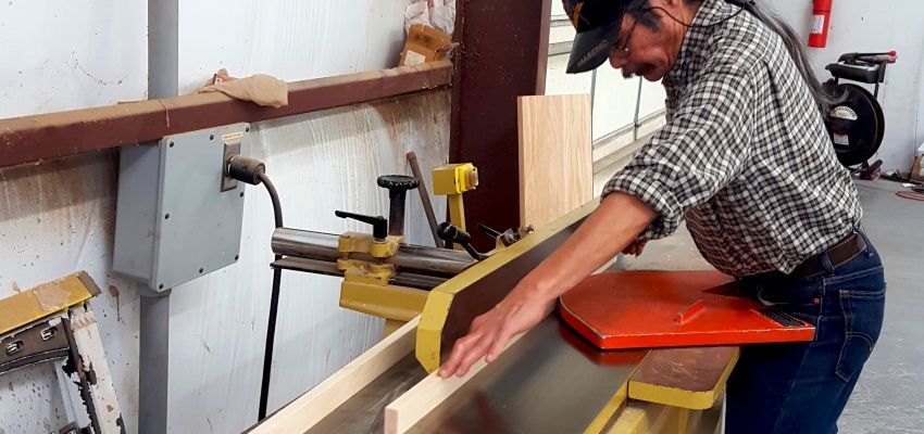 A man in a plaid shirt using a wood cutting machine in a workshop. The machine is yellow and has an orange safety guard.