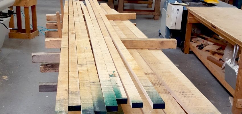 Wooden beams stacked in a workshop. Some beam ends are painted with green or black marks, and the setting appears to be a woodworking shop.