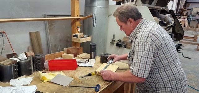 A man in a plaid shirt uses a measuring tool on a wooden board in a workshop. Various tools and materials are on the workbench.