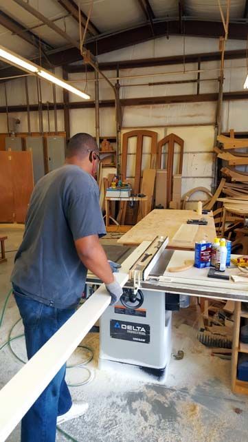 Man in a workshop using a table saw to cut wood. He's wearing safety glasses and gloves. The shop has wood and tools.