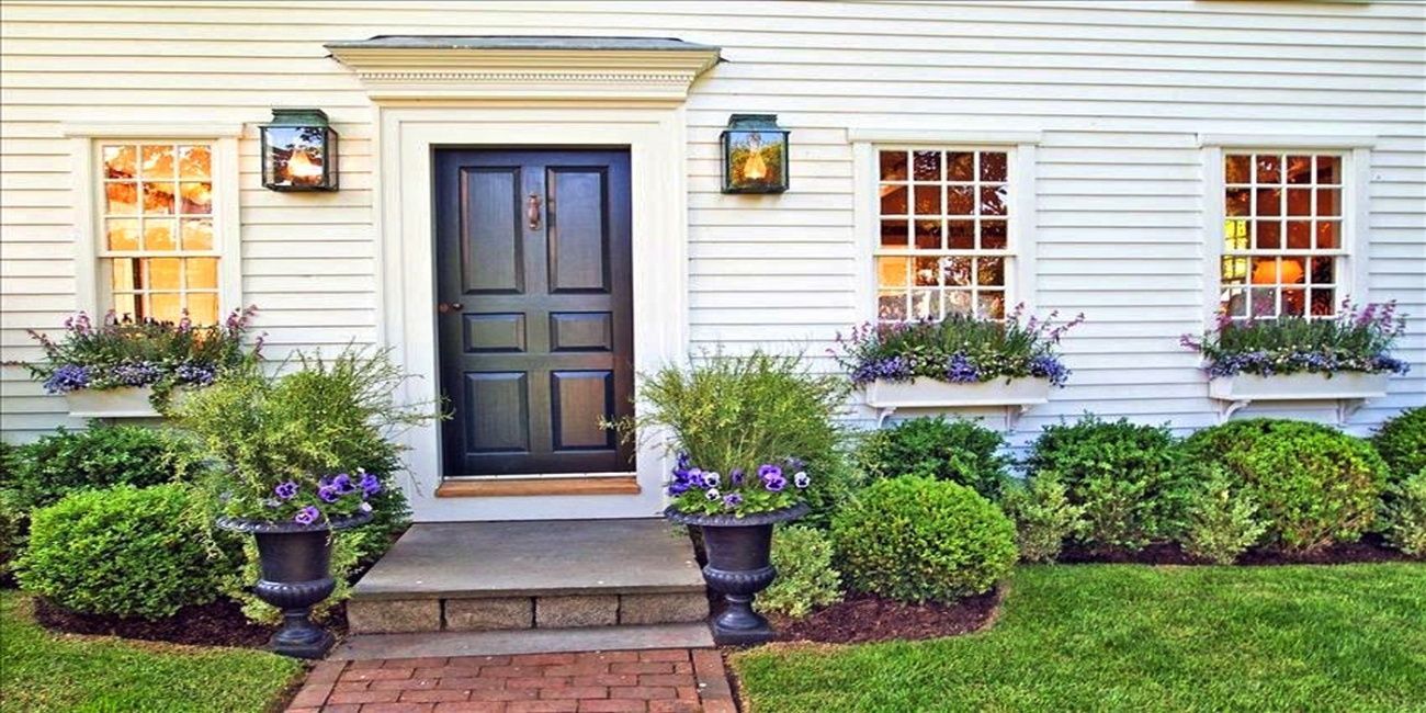 White house with a black door, flanked by windows and lanterns. A brick path and lush landscaping lead to the entrance.