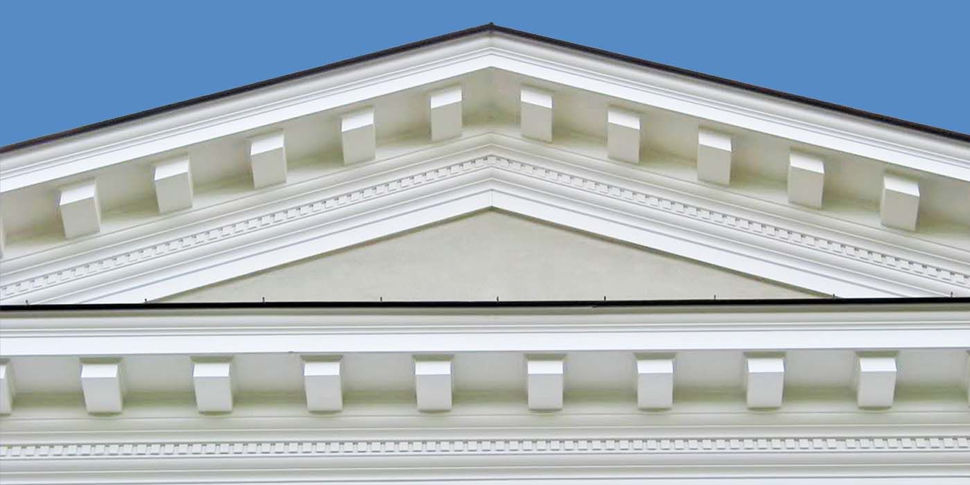 Architectural detail of a white building's pediment, with a blue sky background. Features a triangular shape with decorative blocks.