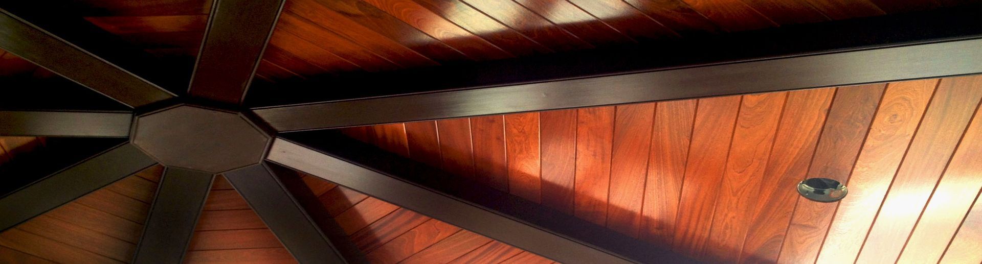 Wooden ceiling with dark beams radiating from a central point. Sunlight casts shadows across the wood panels, and a small light fixture is visible.