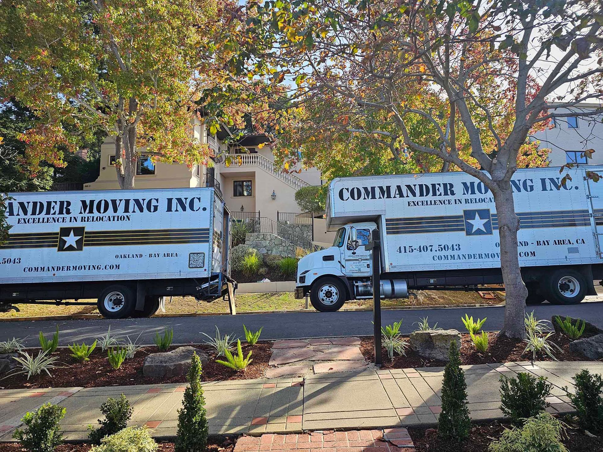 Two Commander Moving Inc. trucks parked on a street with trees and a building in the background.