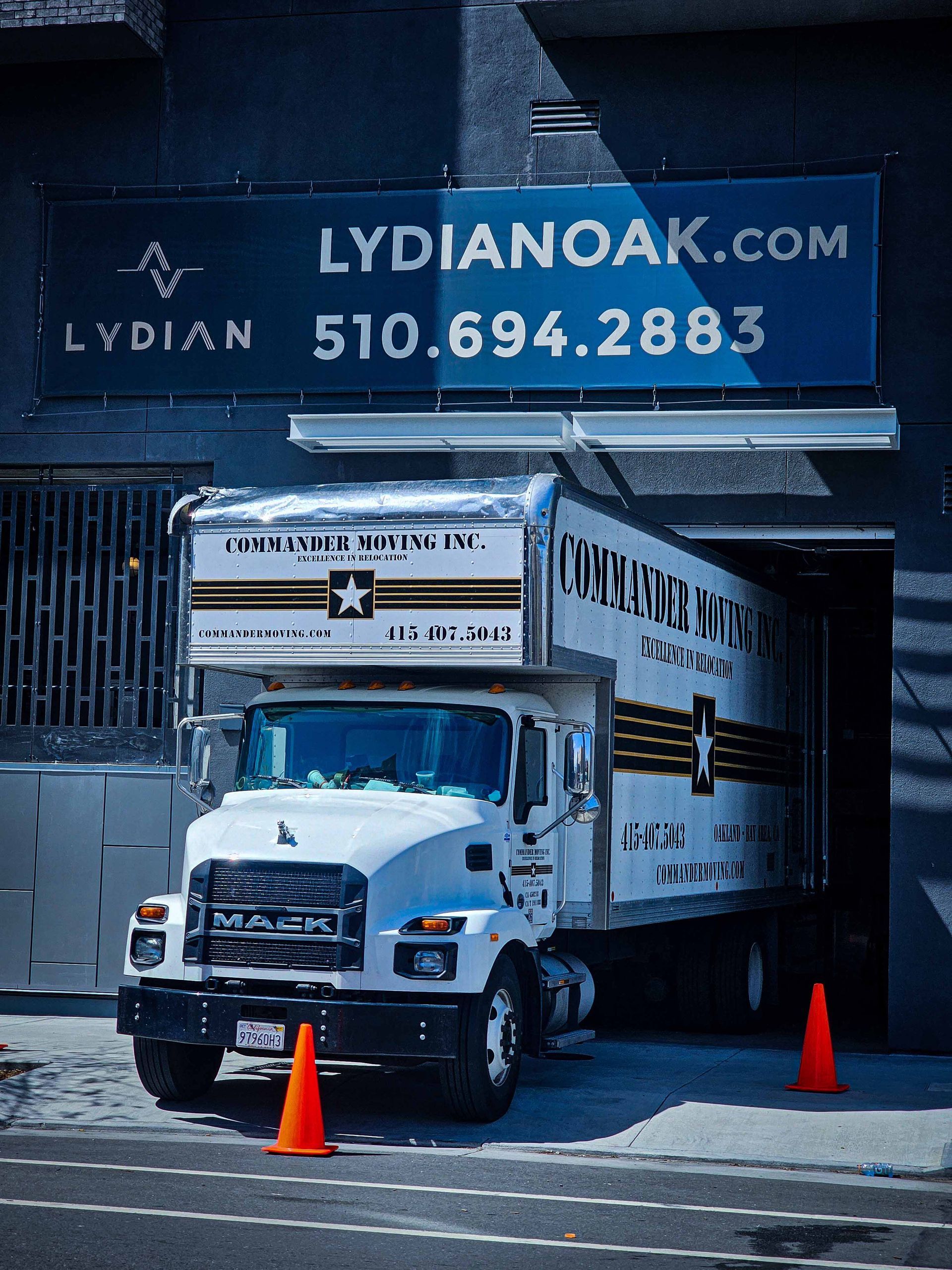 White moving truck in front of a building, sign on top: LYDIANOAK.COM, phone number. Orange cones in front.