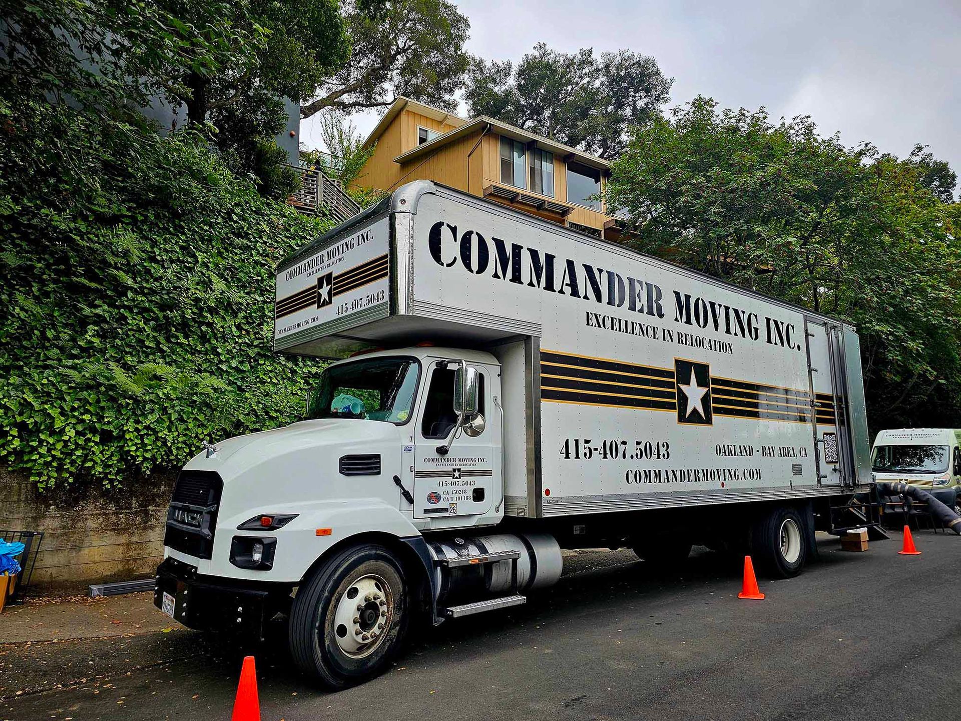 White Commander Moving Co. truck parked on a street, orange cones, hillside, two-story house in background.