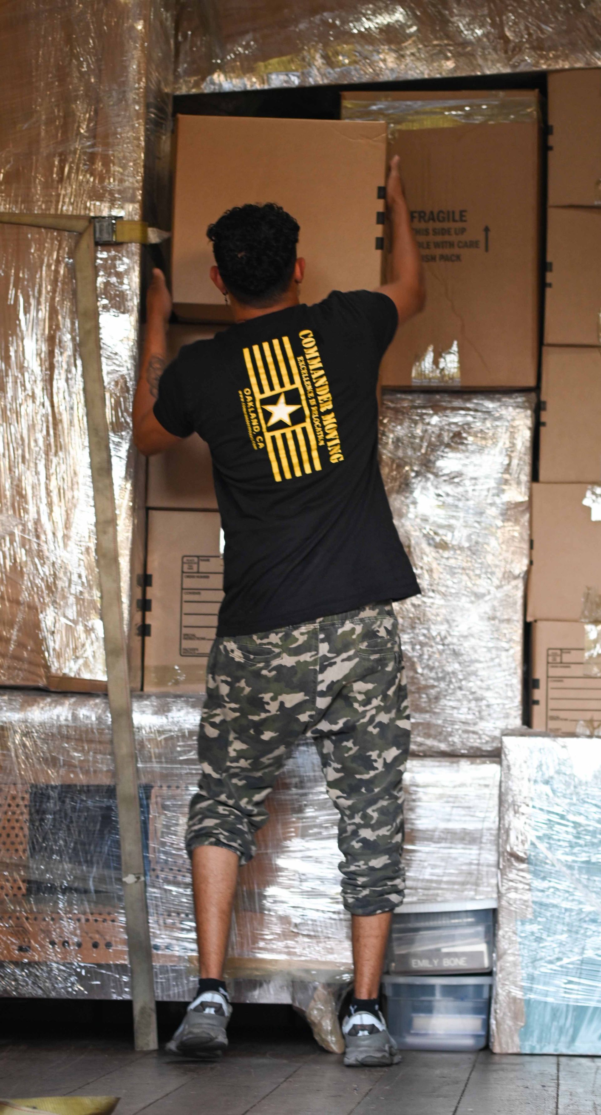 A man in camouflage pants and a black t-shirt stacks boxes in a warehouse.