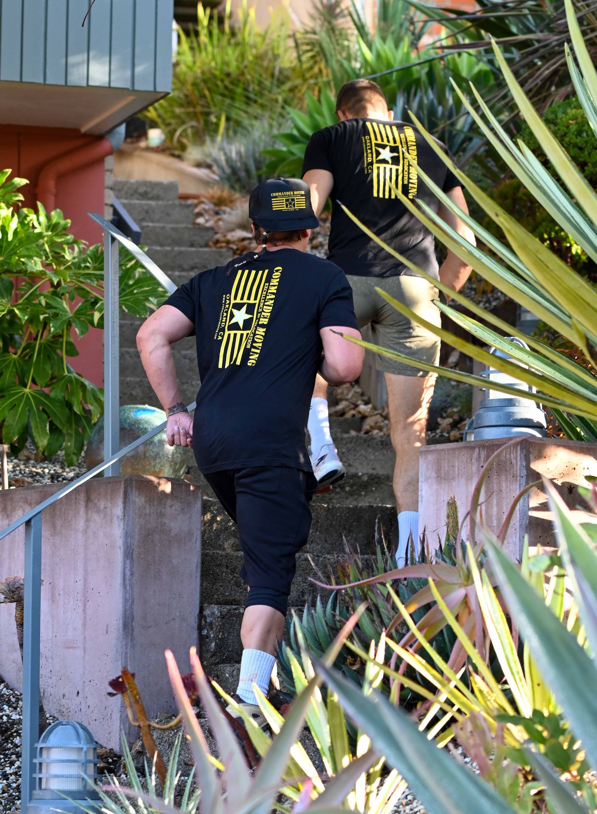 Two men in black shirts run up outdoor stairs, backs to the camera. Yellow logo on shirts. Green plants surround.