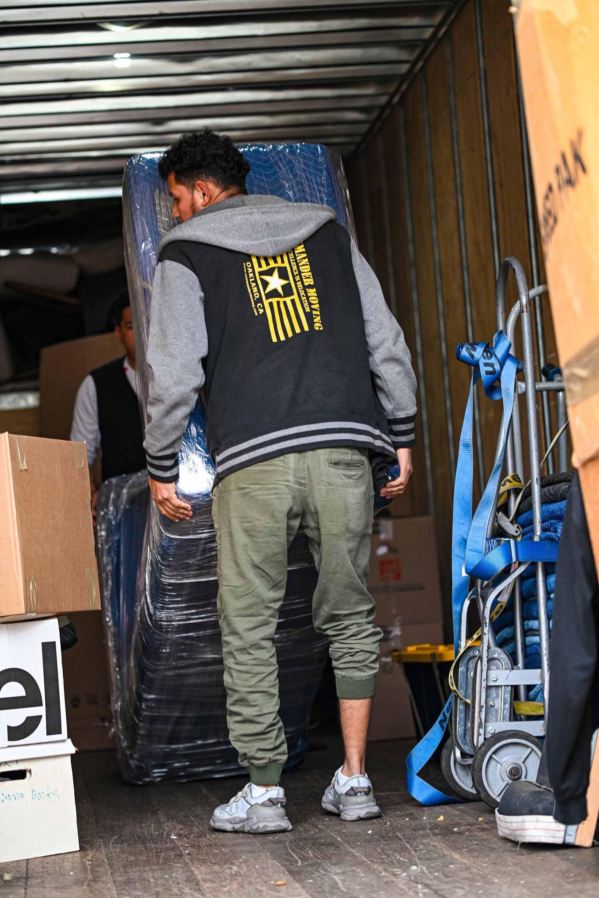 Man in gray jacket and green pants loads a large, wrapped item into a truck, assisted by another person.