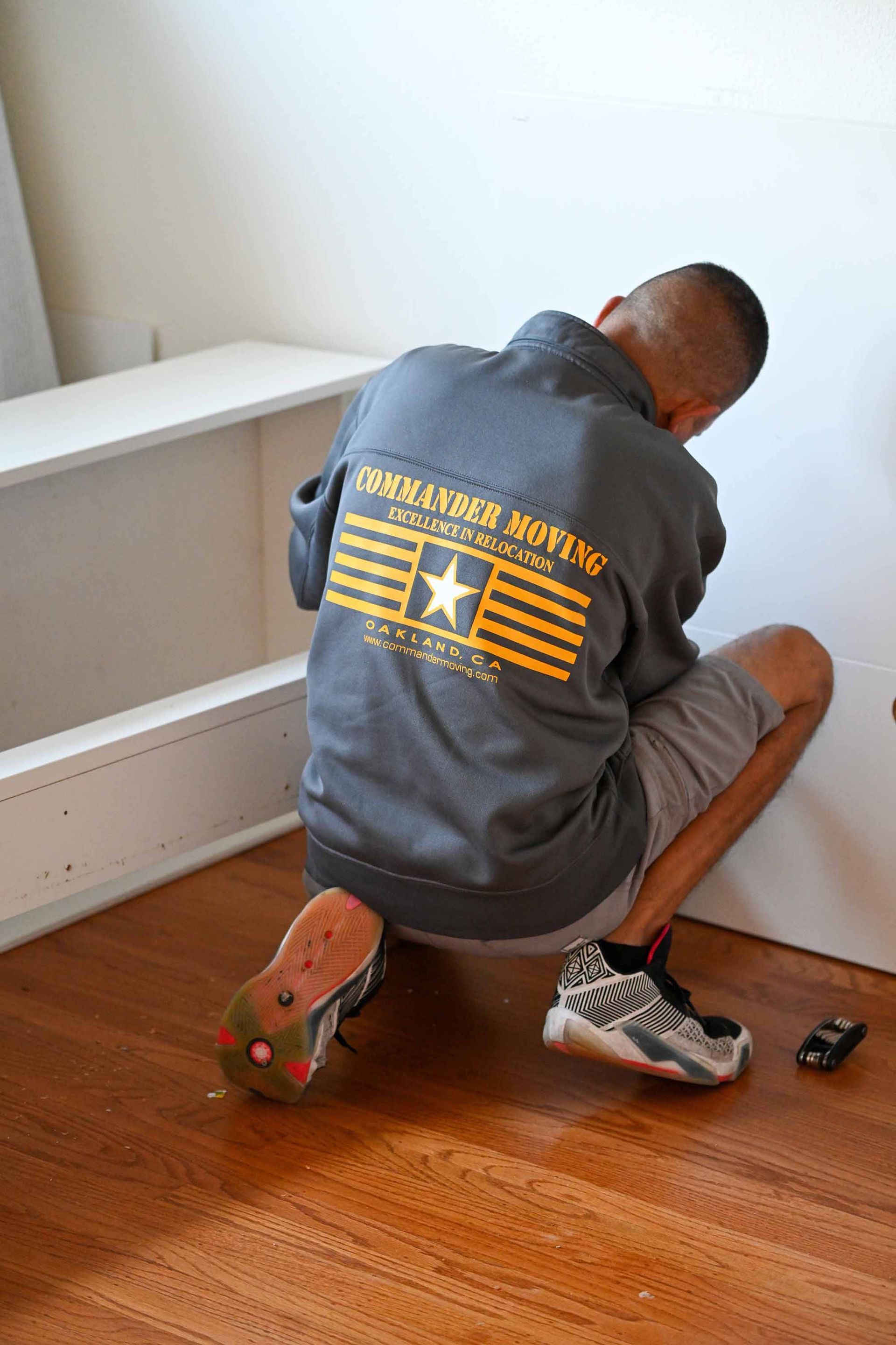 Man in gray sweatshirt crouches, fixing shelf; brown floor, white wall; small toy car visible.