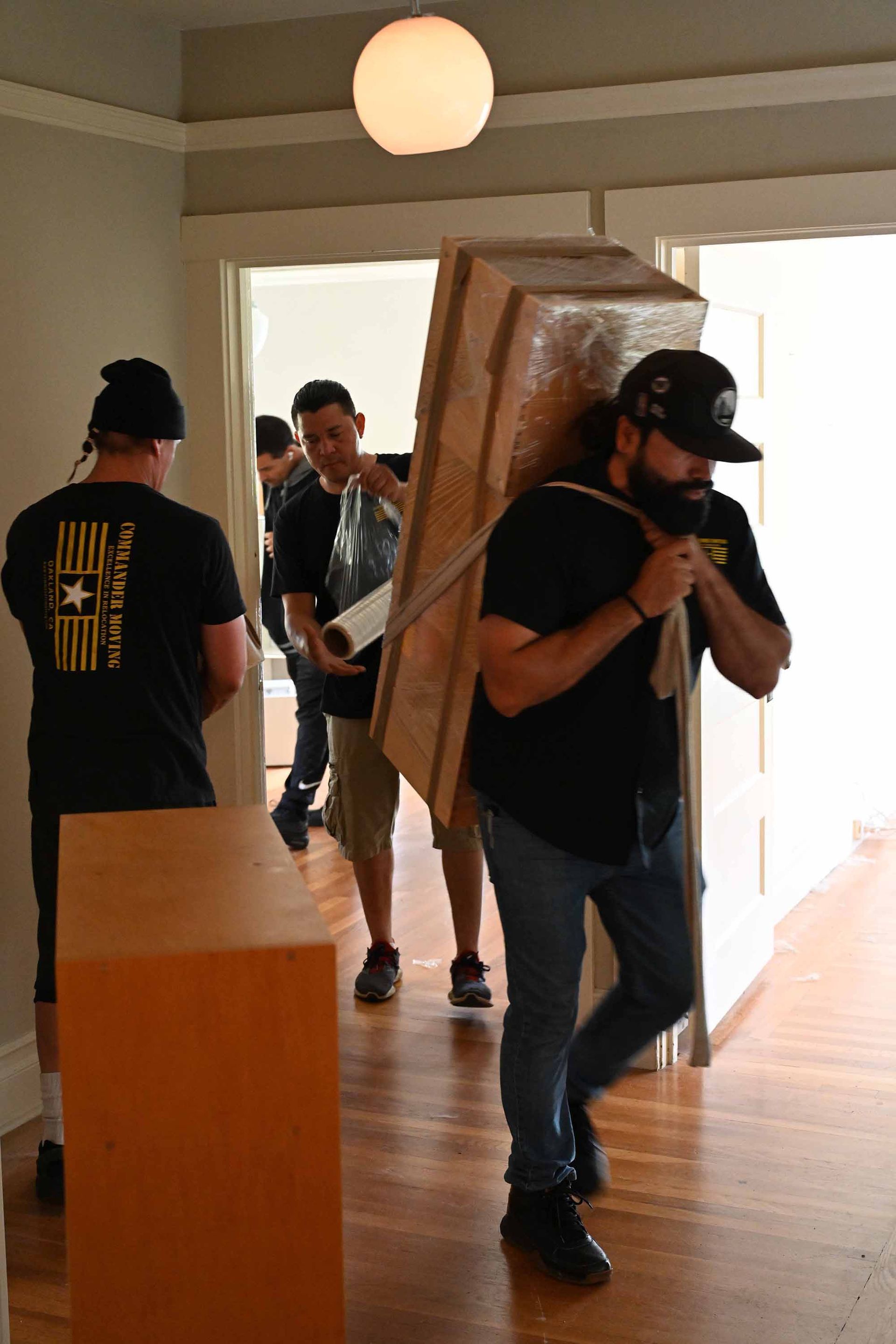 Movers carrying wrapped furniture through a doorway in a house with wood floors.