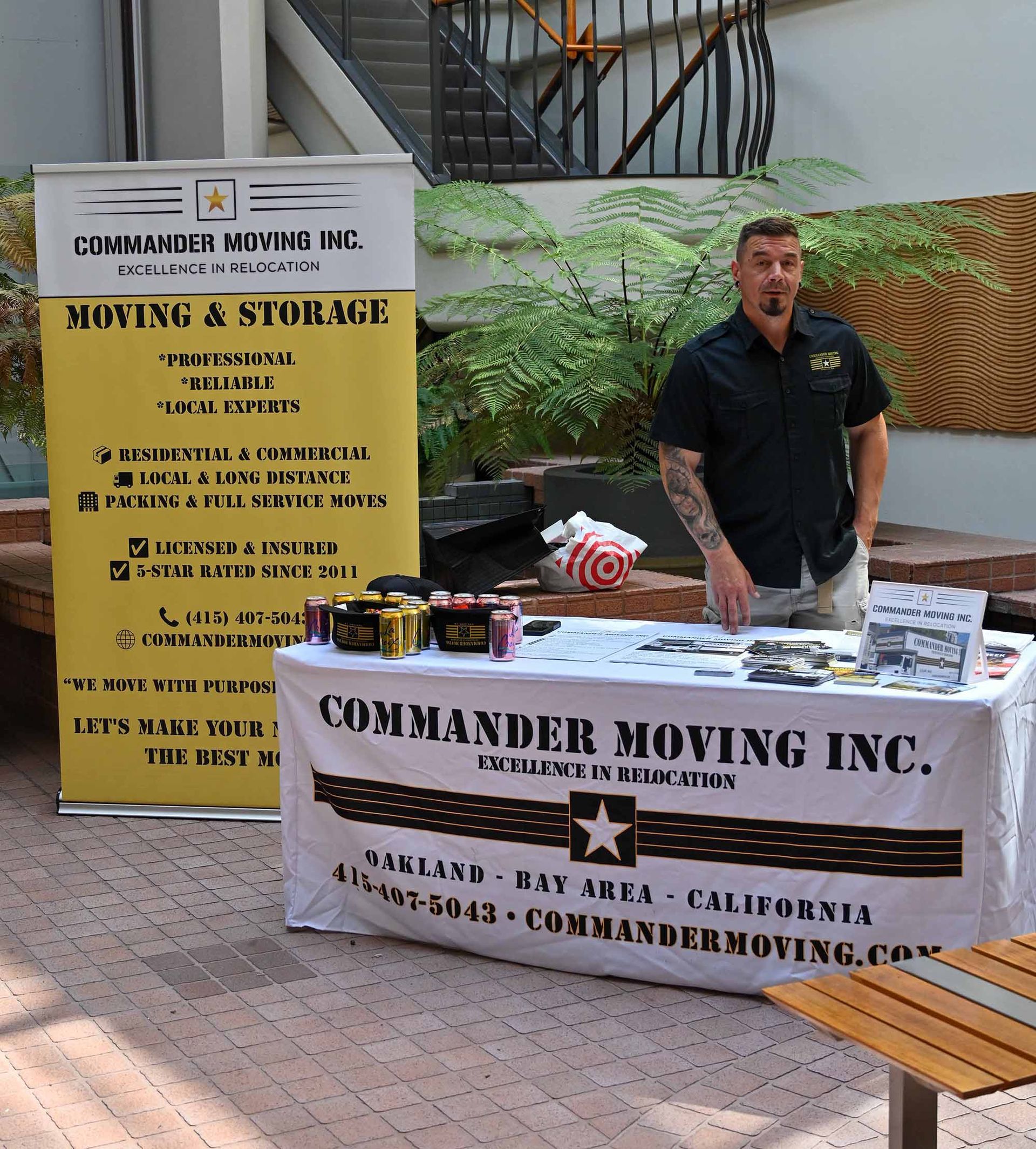 Man stands behind a Commander Moving Inc. table and banner; outdoor setting.