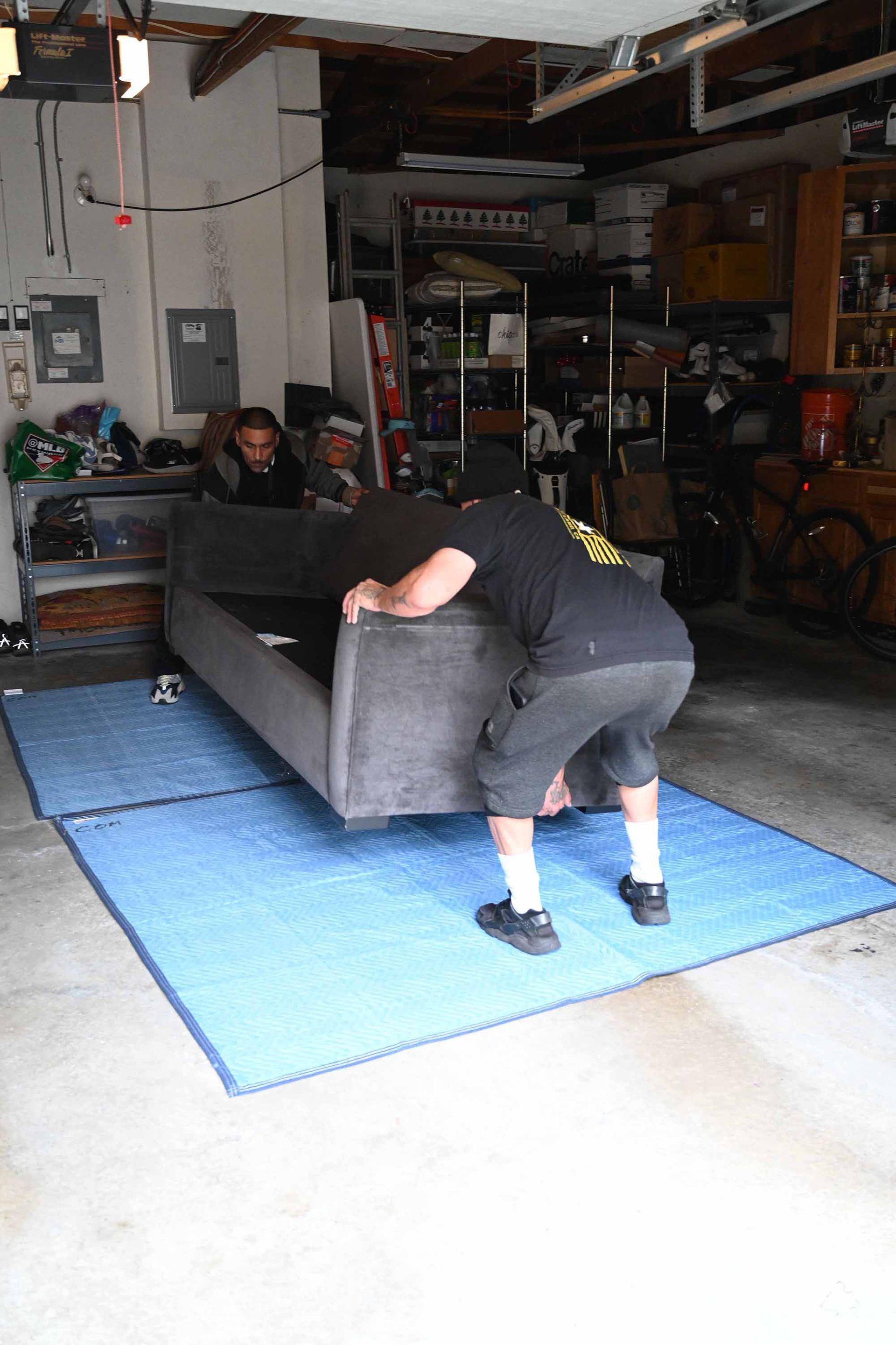 Three people lifting a gray couch in a garage, over blue moving pads.