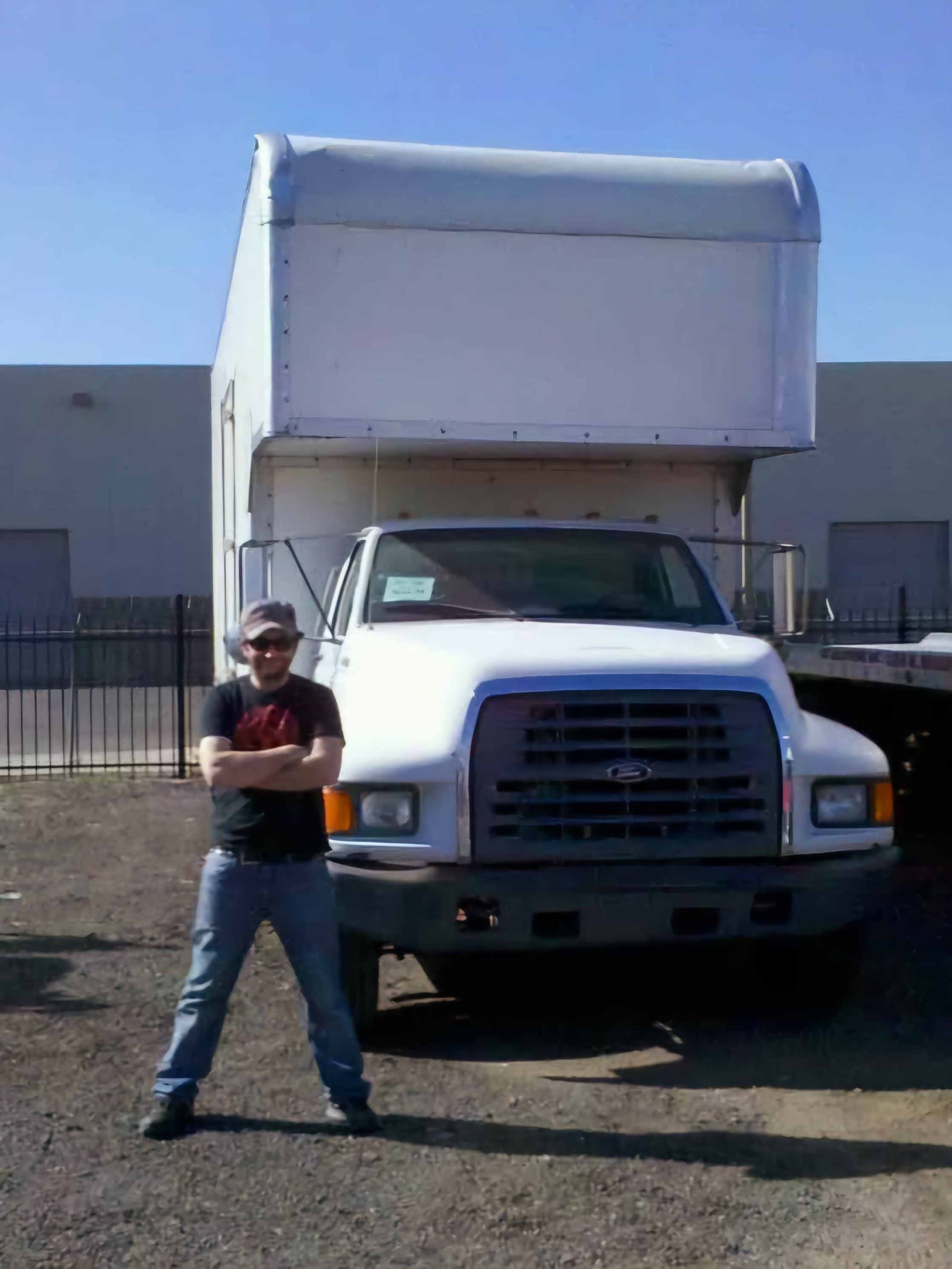 Man standing in front of a white box truck with arms crossed. Outdoors, sunny day.