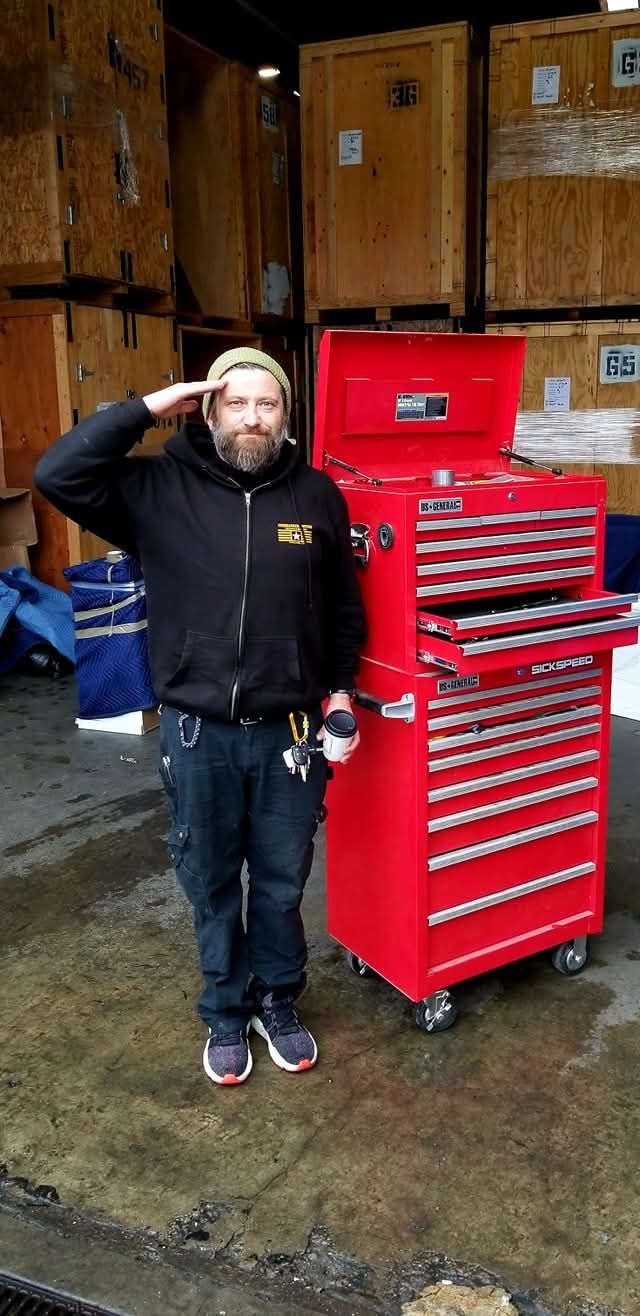 A man in a black jacket salutes next to a red toolbox in a storage area.