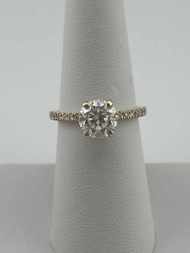 Gold solitaire engagement ring with a round diamond on a white display stand.