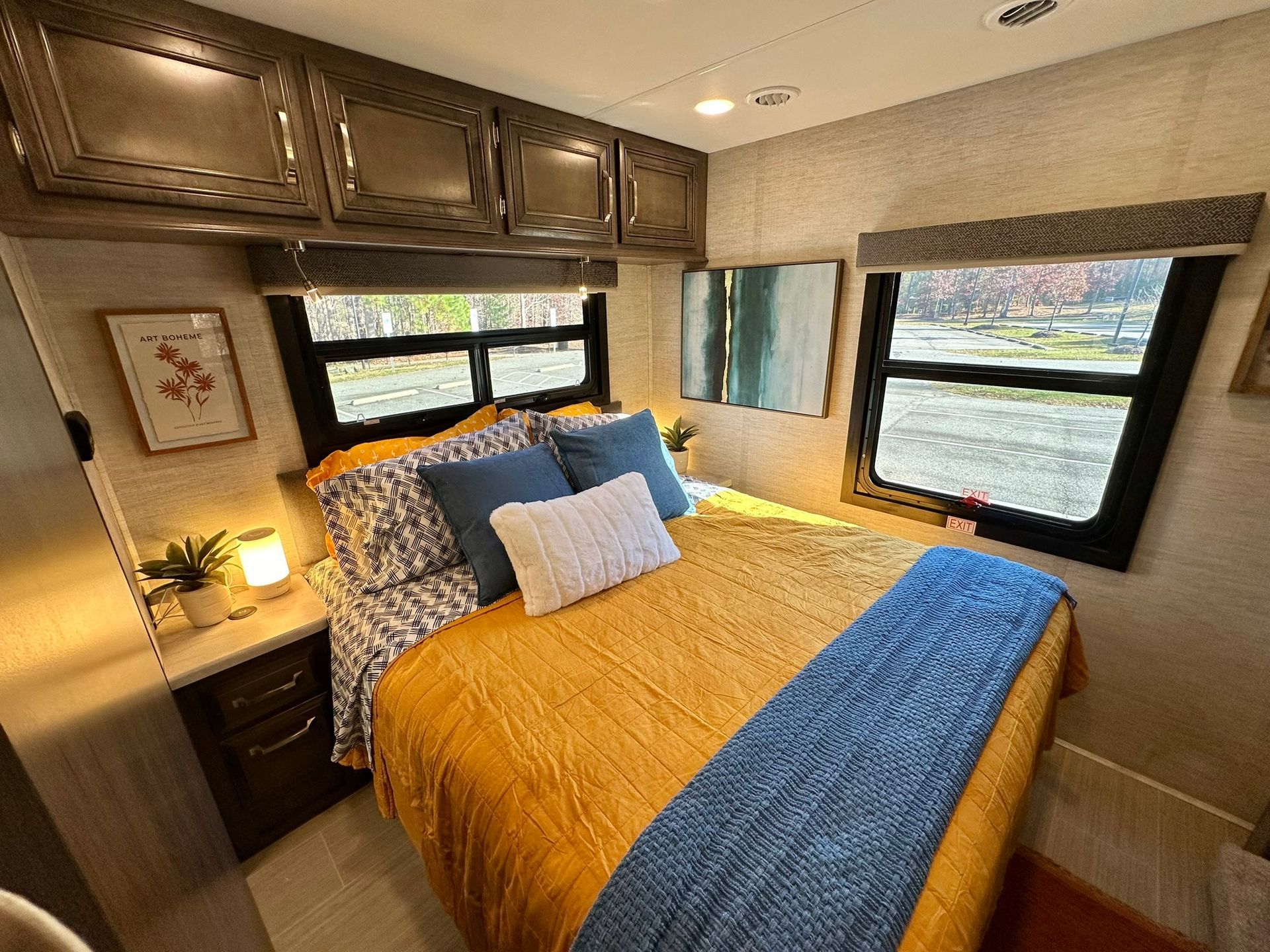 A bedroom in a rv with a bed and two windows.