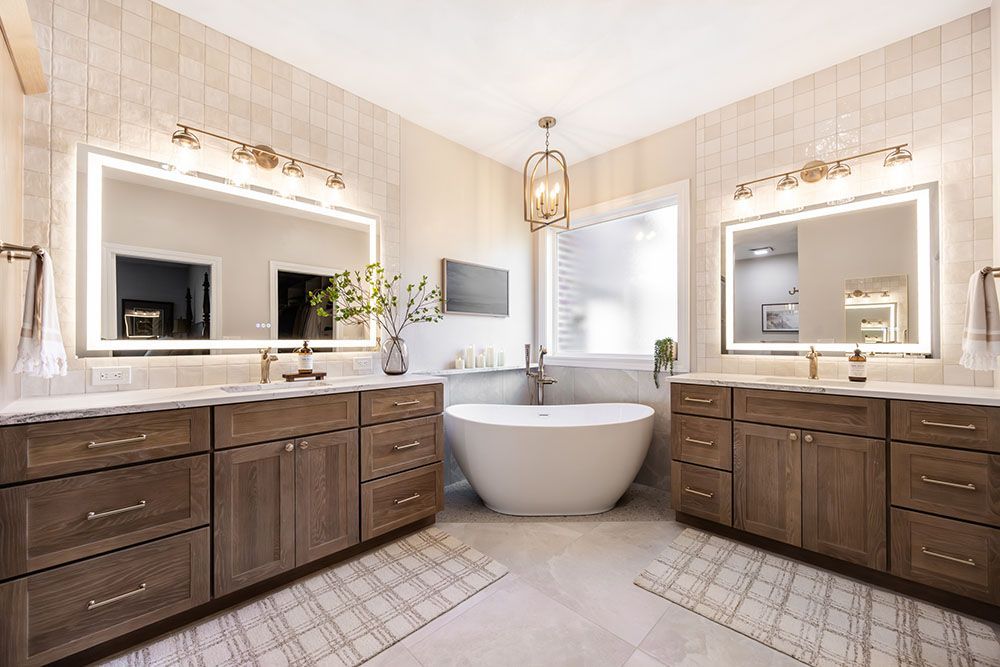 Spacious modern bathroom featuring dual wood vanities with illuminated mirrors, tiled walls, and a central soaking tub.