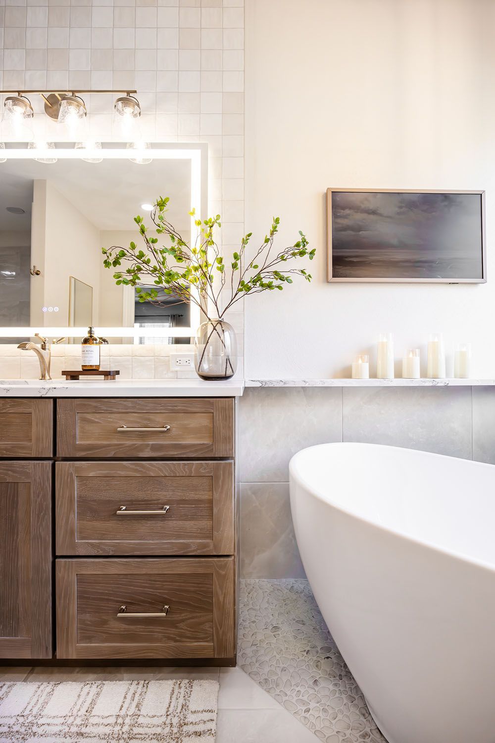 Modern bathroom with wood vanity, white freestanding bathtub, large lighted mirror, and minimalist decor.