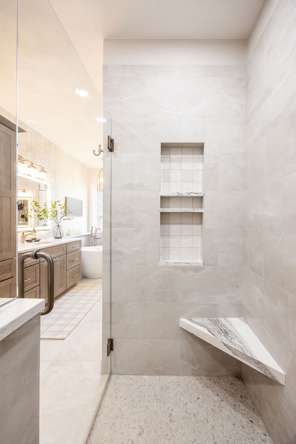 A glass-enclosed shower with light gray tiles, a built-in recessed shelf, and a corner marble bench seat.