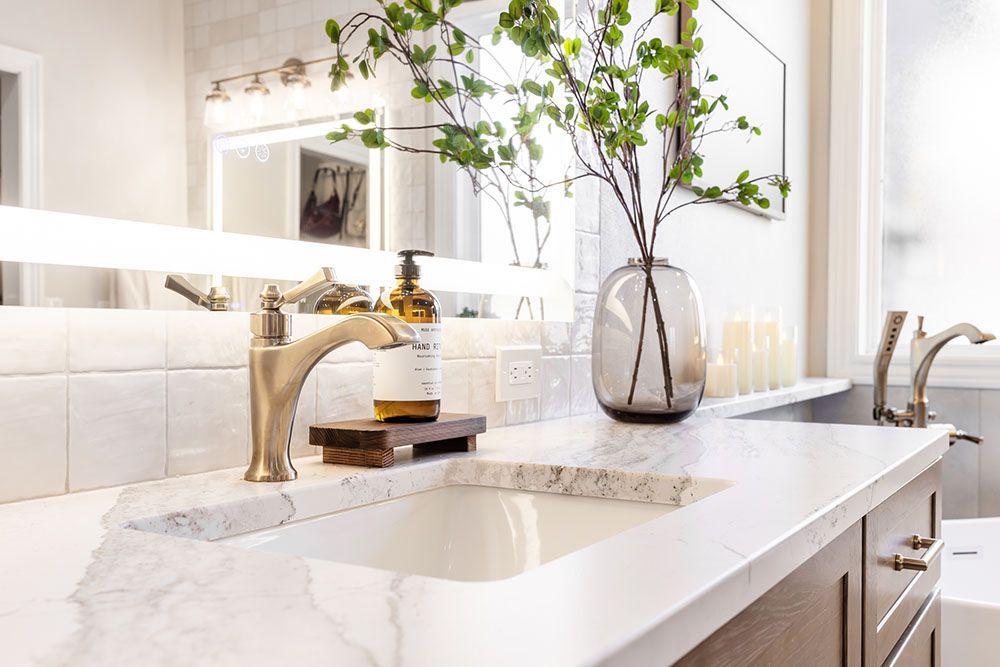 A modern bathroom vanity with a white marble countertop, a brass faucet, a soap dispenser, and a vase with green branches.