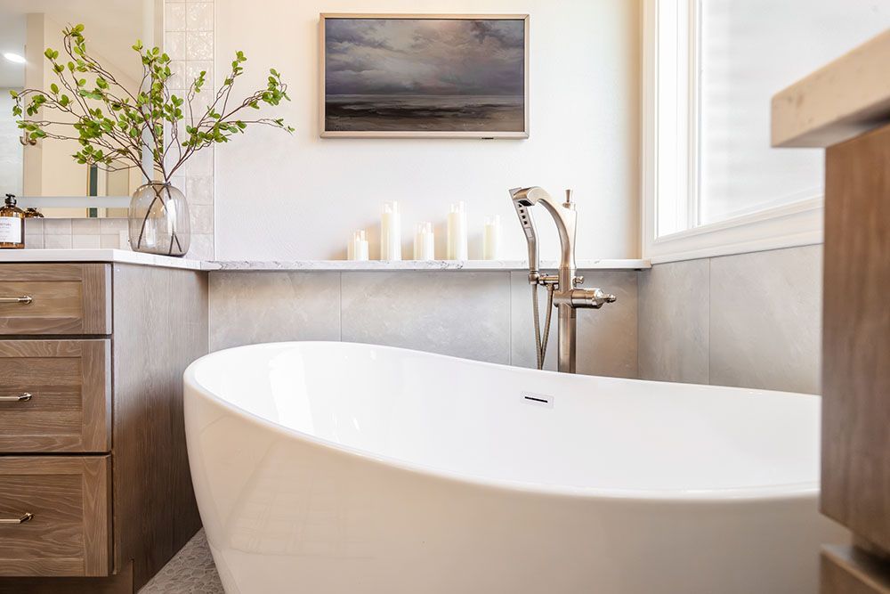 A white freestanding soaking tub next to a wooden vanity with a vase of greenery and candles on a marble ledge.