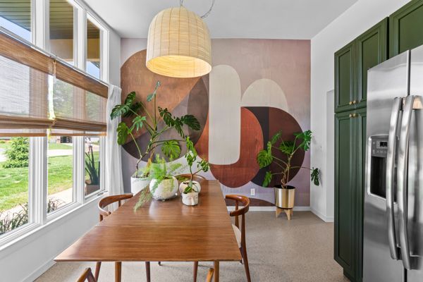 Modern dining room with wooden table, large windows, potted plants, abstract wall art, and green kitchen cabinets