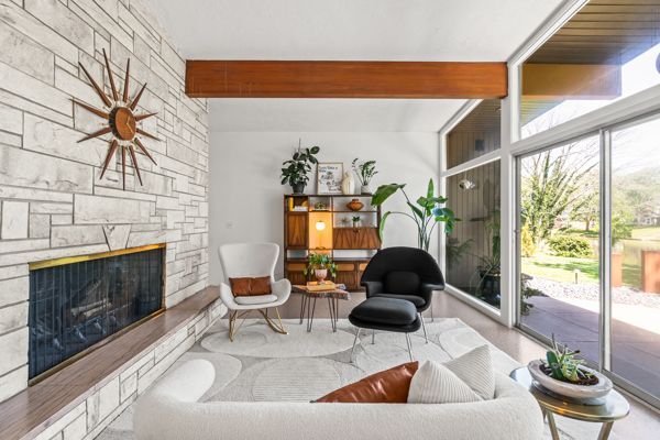 Modern living room with stone fireplace, white chairs, wood accents, and floor-to-ceiling windows overlooking greenery
