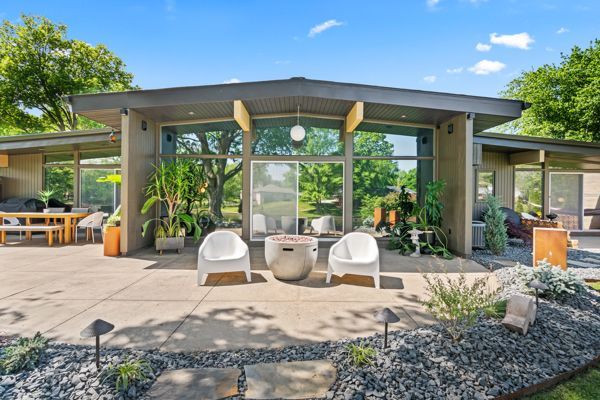 Modern house patio with white chairs, glass doors, and lush greenery under a bright blue sky