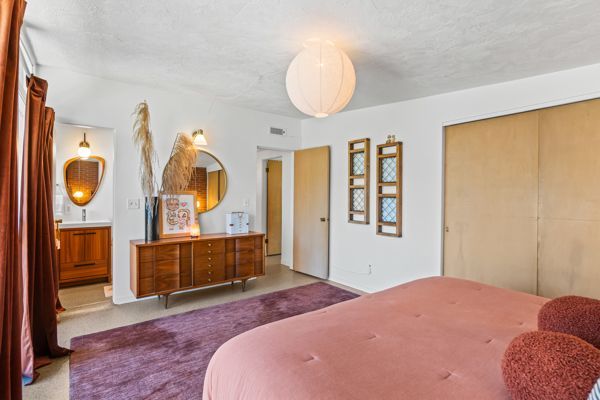 Bright bedroom with pink bedspread, purple rug, wooden dresser, mirrors, and a large window blind.
