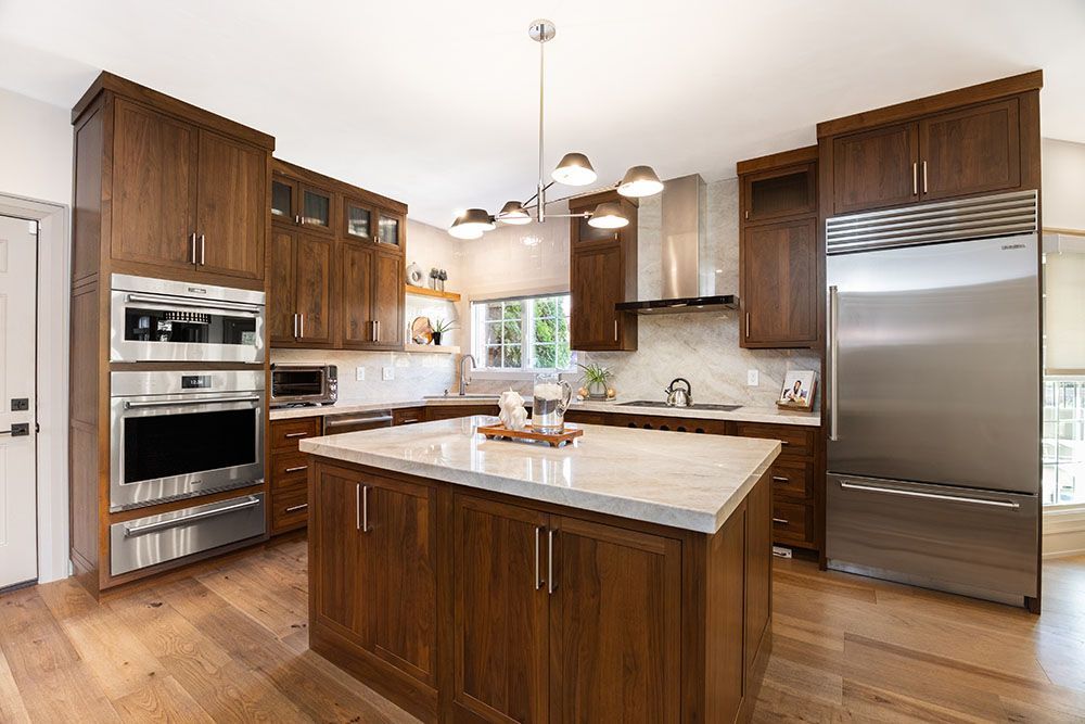 A modern kitchen featuring rich walnut cabinets, light countertops, a central island, and stainless steel appliances.
