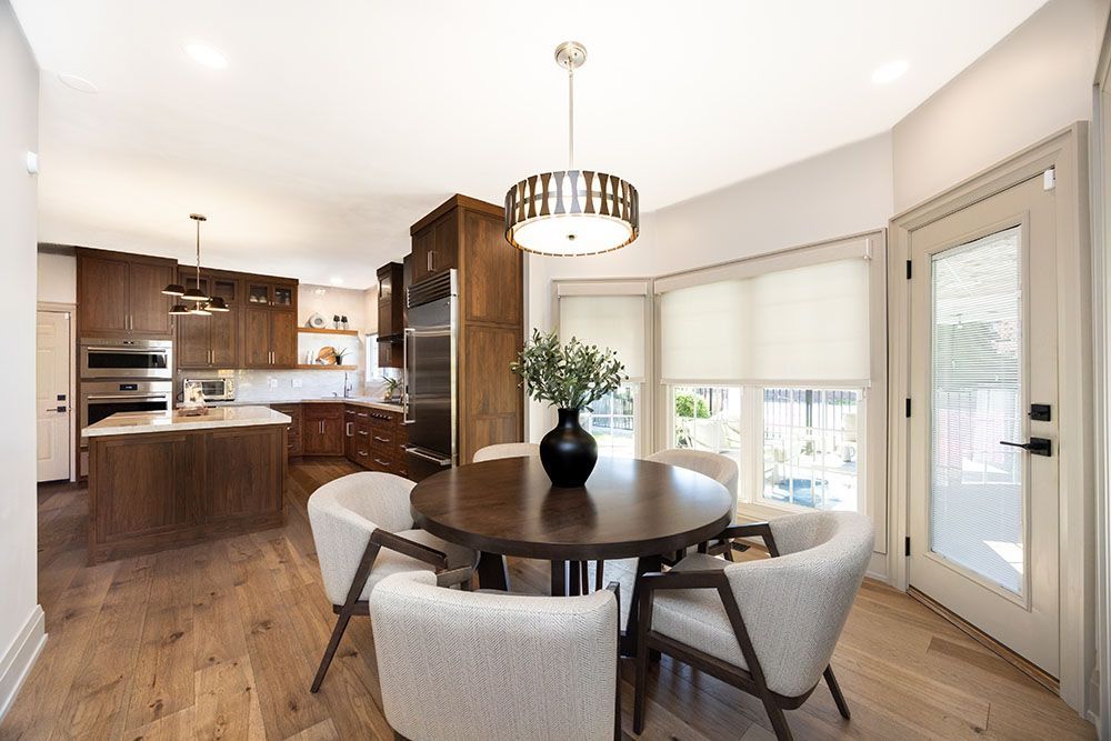 A modern dining area featuring a round wooden table, four light-colored chairs, and a pendant light, adjacent to a kitchen.
