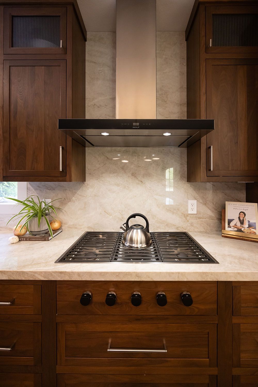 A stainless steel teakettle sits on a gas stovetop in a kitchen with dark wooden cabinets and a marble backsplash.